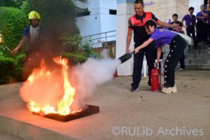 ภาพนางสาวชนิกานต์ บรรจมาตย์ ตัวแทนฝ่ายห้องสมุดวิทยาเขตบางนา ดับเพลิงโดยใช้ถังดับเพลิง เพื่อดับเพลิงที่กำลังลุกไหม้น้ำมัน